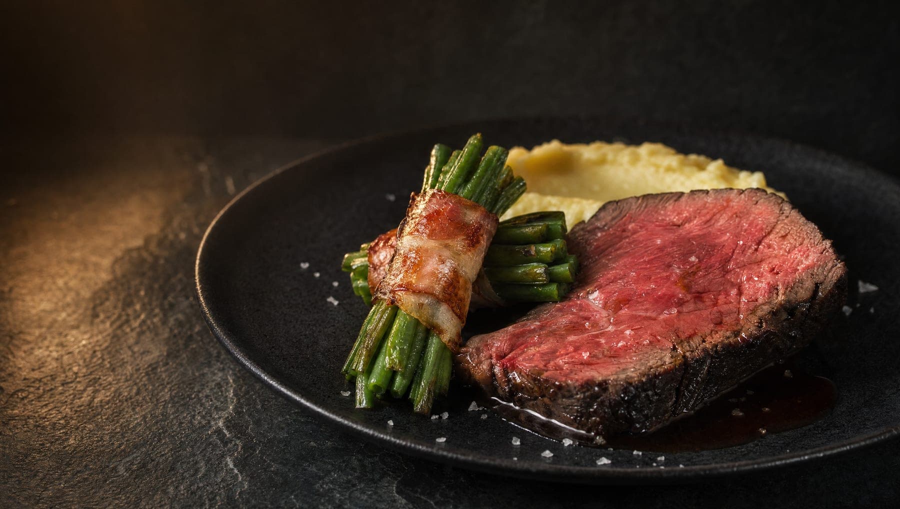Rosa gebratenes Rinderfilet mit Speckbohnen und Kartoffelpüree auf dunklem Teller, warmes Seitenlicht.